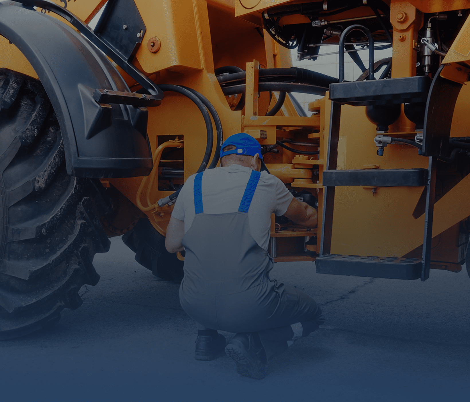 A worker in blue overalls repairing heavy machinery outdoors.