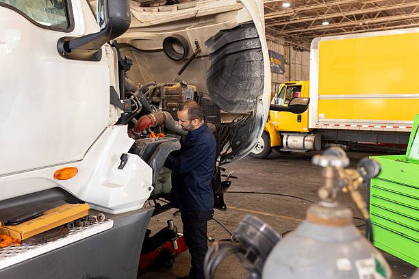 A male truck mechanic works on a large vehicle's engine in a well-lit workshop, surrounded by tools, equipment, and multiple trucks in the background.