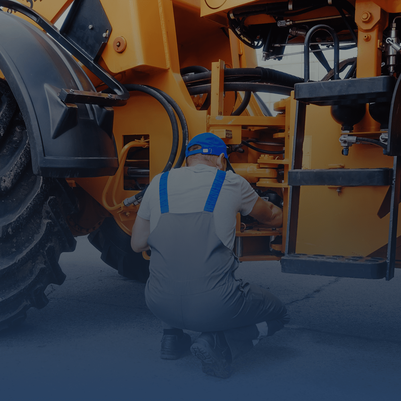 A worker in blue overalls repairing heavy machinery outdoors.